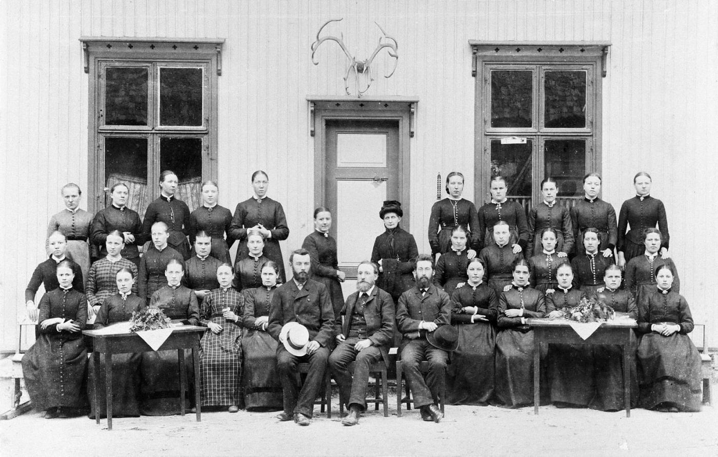 Gruppebilde, svart-kvitt, jenter og nokre få menn i dress. 