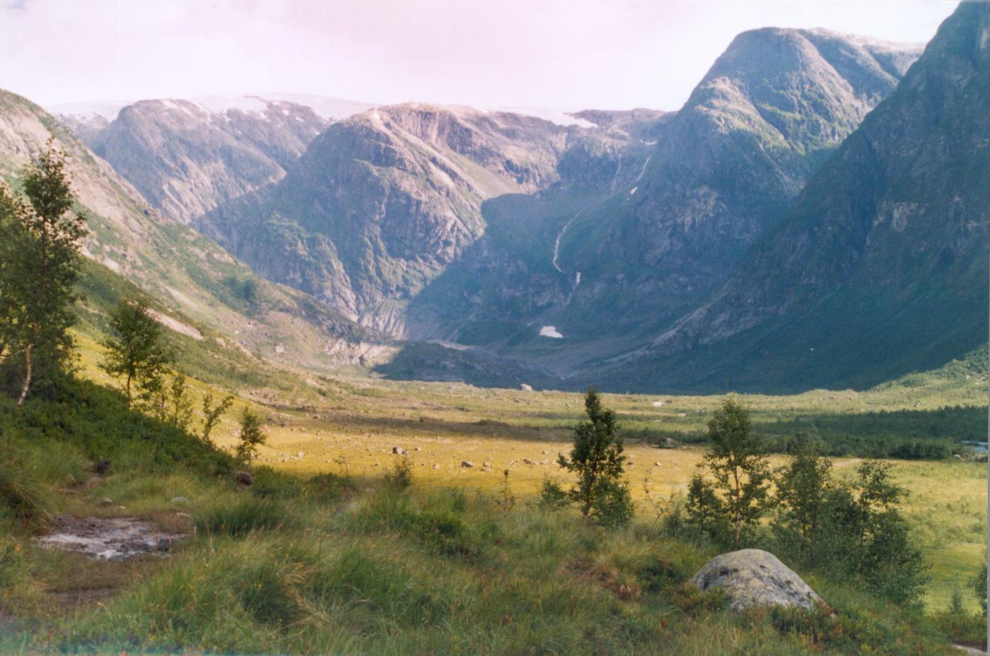 Utsyn nordover Austerdalen. Nærast dannar ei vid slette dalbotnen, lengst inne, under snøfonna, svingar dalen i vestleg retning. Nedste enden av Austerdalsbreen er såvidt synleg rett til venstre for snøfonna. Minnevarden står på fjellryggen der dalen svingar.