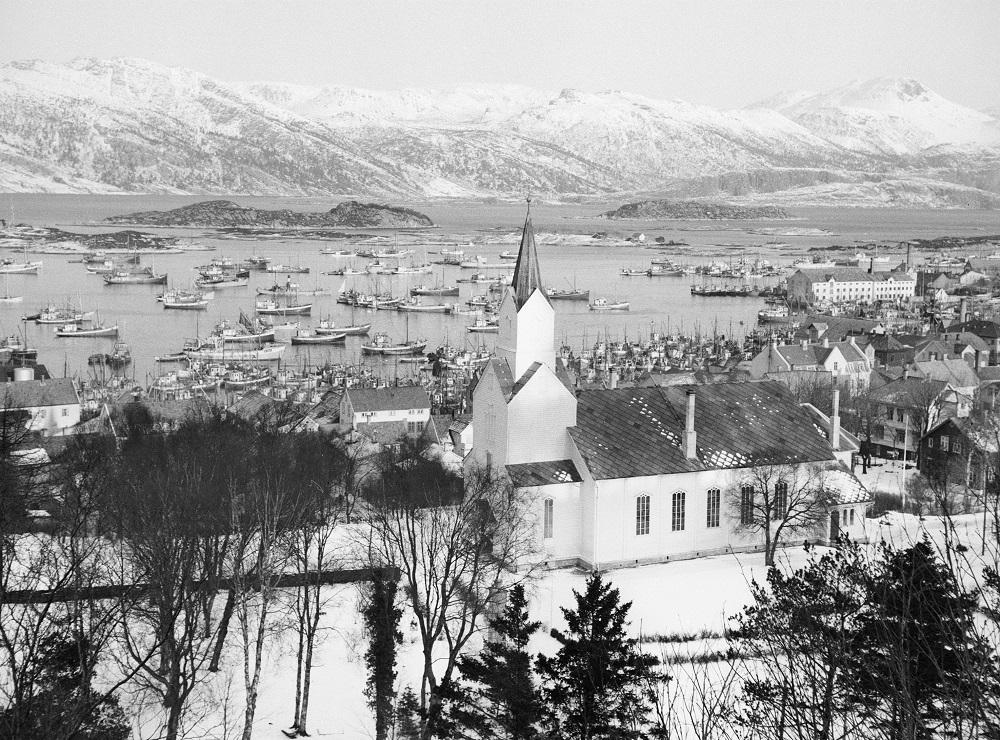 Utsyn frå Likkjeåsen og nord-austover, med kyrkja i framgrunnen, byen og fjella i nordaust. Fiskarflåten ligg i hamn, februar 1953.