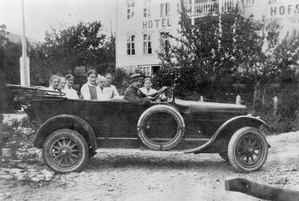 Dette er den første turbilen i Indre Sogn Automobilselskap A/S,  ein Chandler med sju seter, kjøpt i Bergen i juni 1916. Sjåføren er Anton Andersen som og var bilmekanikar. Han var såkalla garantisjåfør den første tida. Ingen andre kunne køyra bil i Sogndal. Passasjerane er truleg jenter frå Sogndal. Fartsgrensa var 30 km/t, og selskapet måtte stilla garanti for mogeleg skade før køyringa tok til, sjølv om det ikkje var sjåføren si skuld.

 