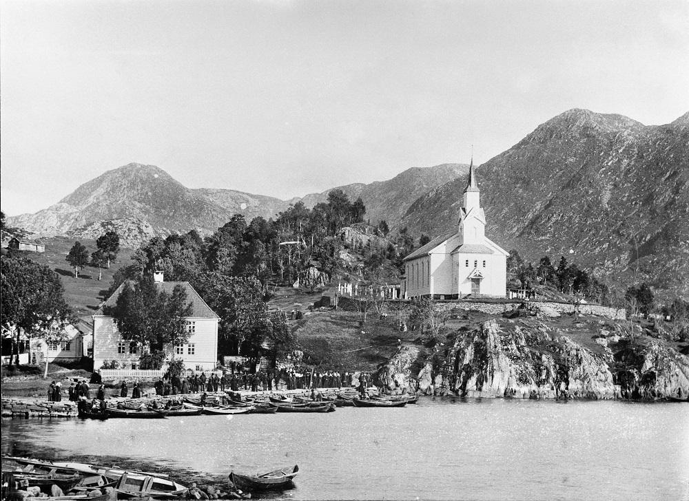 <p>Gulen Kyrkje og prestegard i Eivindvik med kyrkjelyden og helgepynta folk p&aring; kaia. Nesten alle kom sj&oslash;vegen. Kyrkja vart bygd i 1863. Vegen langs stranda kom fyrst i 1923.</p>