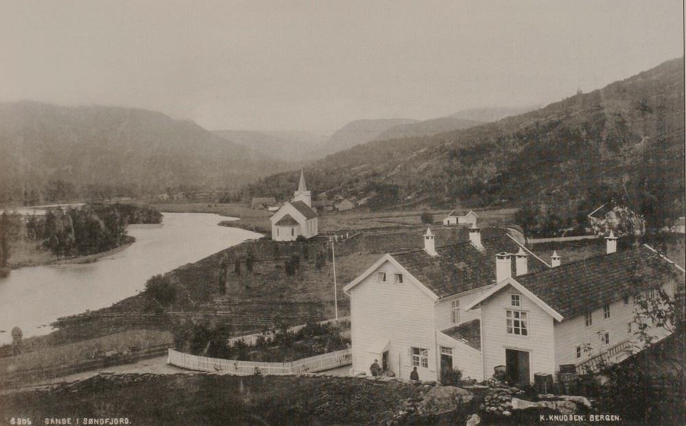 Framme i biletet ser vi Sivertsens hotell. Lyder Sivertsen eigde og dreiv hotellet i 1905. Her var både landhandel og skysstasjon og postopneri i tillegg til hotelldrifta. <br />
Lenger bak ser vi Sande kyrkje, bygd i 1864 av byggmeister John Saftland, arkitekt: J. Lindstrøm.
