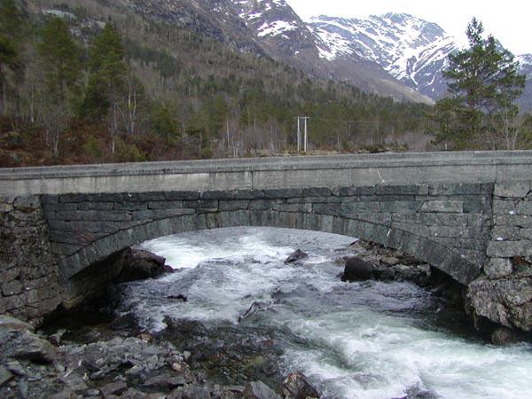 Brua sett frå sør mot Hope. Det er støypt betongrekkverk på båe sider av sjølve brua og fyllingane inn mot terrenget.
