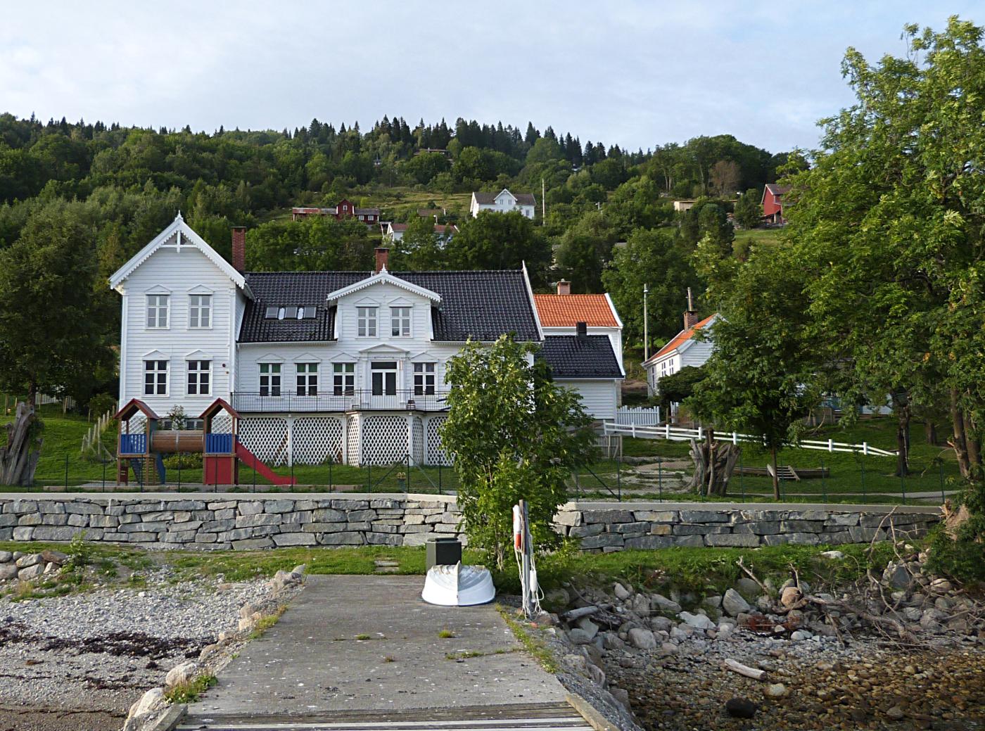 Bilete av kvit gamal trebygning, teke frå ei brygge. 