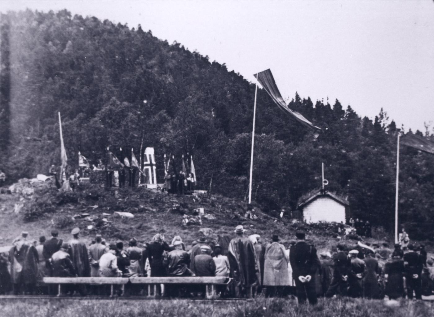 Frå avdukingshøgtida ved ferjestaden Våge søndag, 15. august 1943. Bautasteinen er sveipt i det norske flagget og omkransa av flagg og banner. Heimestyrkar øydela minnesmerket i juni 1945.