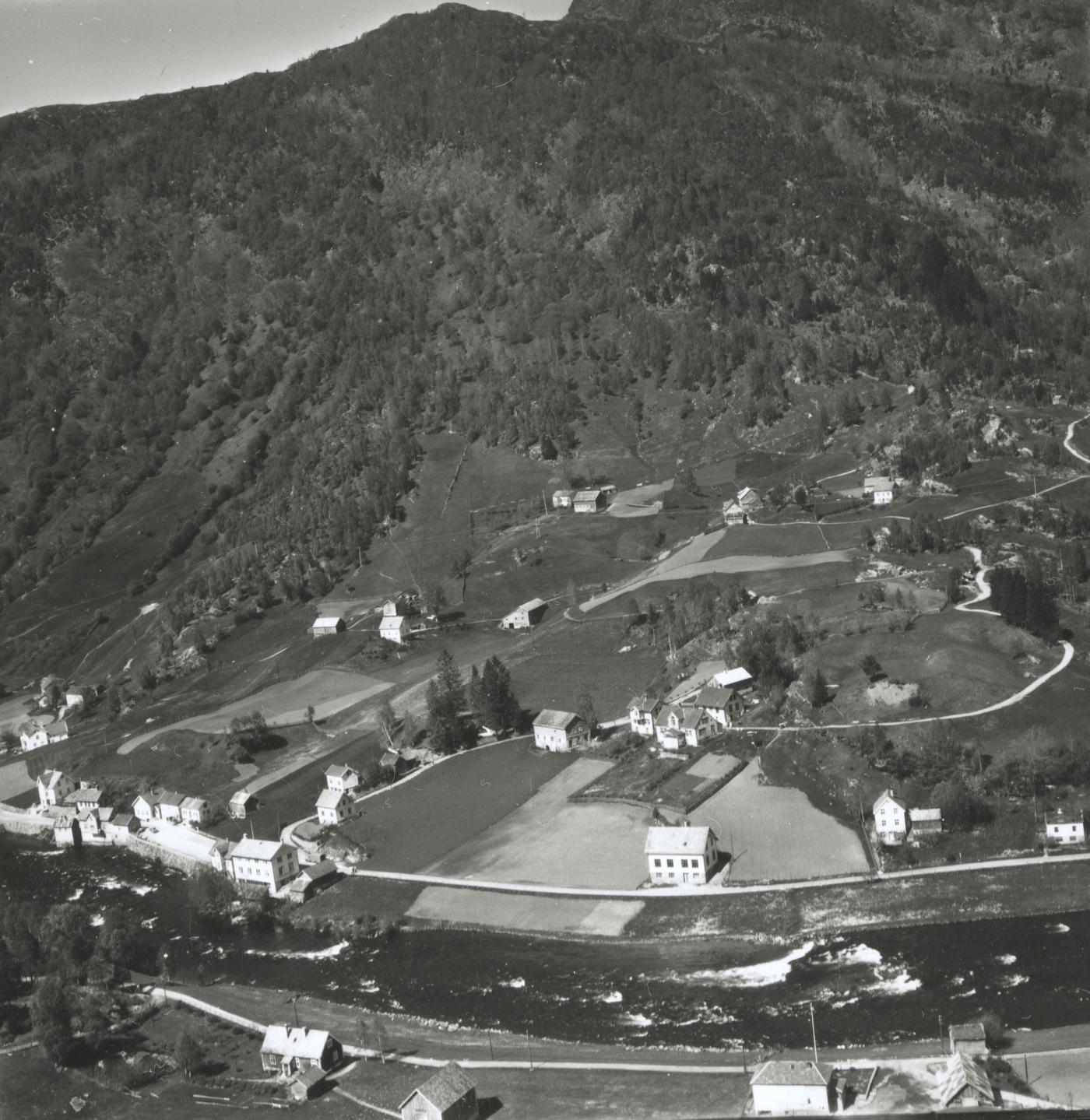 Flyfoto av Skagen i 1956. Oppe til venstre ligg bruk 1, deretter bruk 2/4 og bruk 3 oppe til høgre. Midt til venstre ligg bruk 5, Nedre Skagen. Det var frå denne eigedommen at eigedommane nede ved elva vart frådelte. Desse var då utgangspunktet for dei sentrumsfunksjonane som Skagen hadde før sentrum vart flytta der det ligg i dag. Det gamle kommunehuset ligg øvst i den tette husklynga. Oppafor der ser vi framleis restane etter den gamle brua på Sande.