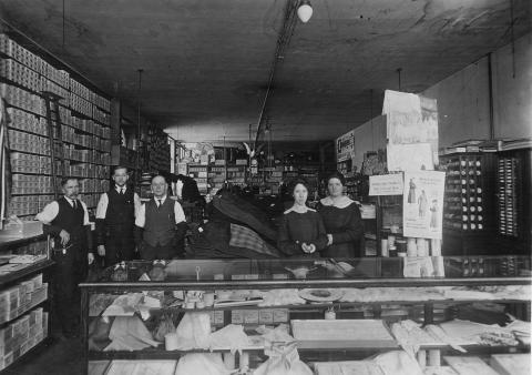 Interiør frå butikken til Andrew Opheim. Han står lengst til venstre, ved sida av Odd Eide. (Voss folkemuseum)