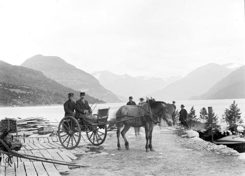 Dampbåten «Faleide» til kai (Postvegbryggja) på Faleide. Ungguten til venstre i skysskjerra er Gustav A. Faleide (f. 1873). Han var son til partseigar i båten, Anders H. Faleide, og bror til styreleiar Hans A. Faleide. Rett bak hesten står ein annan partseigar i båten, Rasmus A. Faleide. Dei andre personane er ukjende. Båten er pynta med 6-7 furebusker og folk er oppkledde, sikkert i høve ei spesiell hending. Biletet er teke før båten fekk overbygg og ein høgare og tunnare skorstein enn den opphavlege.