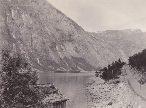 Biletet viser slagskipet til ankers i Simadalsfjorden, ved Vik i Eidfjord kommune. Naustet ligg eit kort stykke utom Stavaneset.