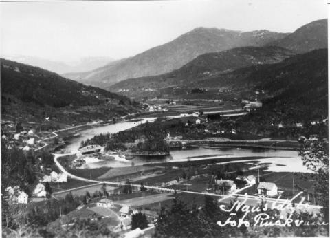 Biletet viser Naustdal og brua over Nausta ved Naustdalsfossen i 1940-åra. Brua, som er ei fagverksbru, vart bygd i 1923. Vegen til Førde går mot høgre biletkant. Det var strid i kommunen om kor brua skulle byggjast. Mange ville ha bru nede ved "strandstedet". 