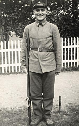 Olav Hjelle (1917-1940). Her er han i uniform heime då han var i "eksisen" i 1938. Det var Olav som sette opp stakittgjerdet i bakgrunnen. Olav var flink skihoppar. Den gongen gjekk dei lange vegar til skirenn, og det var nok difor dei fekk så god "kondis", fortalde Petra Nes Hjelle ved årsskiftet 2002/2003.
