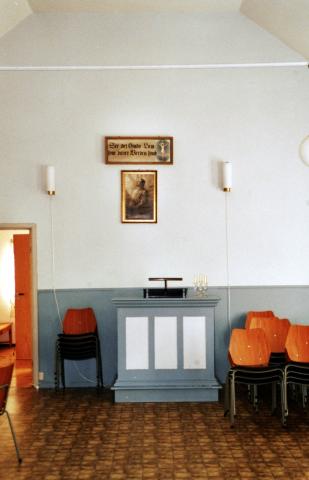 <p>Talarstolen st&aring;r p&aring; den motsette enden av inngangsd&oslash;ra. Dei opphavlege benkene vart skifta ut med lause stolar som lett kan plasserast etter behov. Det innramma bibelordet, med skrivefeil, er: "See det Guds Lam som b&aelig;rer Verden synd."</p>