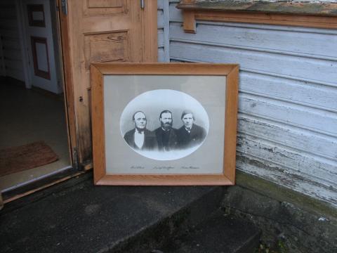 Innramma bilete på veggen i Storsalen (her på inngangstroppa for avfotografering), føregangsmenn for indremisjon og bedehus i Lærdal:  Knut Helland, Ingebrigt Christoffersen og Thomas Thomassen.