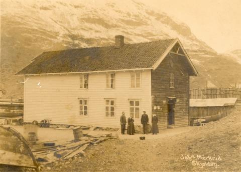 Kaihuset, eller Bryggjehuset, kring 1910. Inngangsdøra var på denne tida på endeveggen opp mot vegen. Personane på biletet er frå venstre: Johannes J. Mørkrid og kona Ingeborg, sonen Kristen, som vart gardbrukar i Fortun og dottera Elina som utvandra til Amerika, Nord Dakota. Stolpeveggen til høgre høyrer til Sogns Iskompagni sitt anlegg som var i drift frå 1893. 
