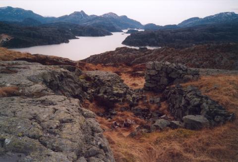 Rest av grunnmur frå sjølve vakthytta på Hugøyknolten. Vi ser sør i Hagefjorden. Ingen bygningar står att, men murar og gammal piggtråd fortel dyster historie.