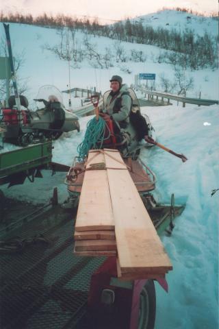 Materiellet til ny kledning på stolpeskuret i Horndalen vart frakta inn med helikopter hausten 1999 men ein fekk for lite kledningsbord. Resten av kledninga vart difor køyrt  inn med snøscooter våren 2001. Lærdal Røde Kors Hjelpekorps tok oppdraget som ei øving og for å bli kjende i området. Her ser vi Franz Henriksen i hjelpekorpset har fått materialen på kjelken og er klar til start.