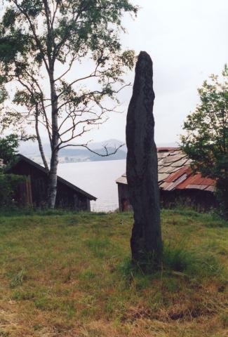 Bautasteinen står no på flat mark rett oppfor ei naustrekkje.
