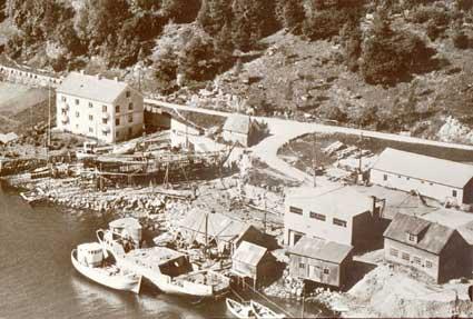 Løland Motorverkstad ein sommardag i 1956. Husa ved kaia er frå venstre: saghuset, eit lagerskur og motorhuset. Til høgre det gamle verkstadbygget med bustad og seinare matsal i andre høgda. I halvtekket på oppsida vart traller o.l. laga. Huset brann ned i desember 1992. Til venstre smia der m.a. lufthammaren stod. Snikkarverkstad i andre høgda. Vegvesenet sitt lagerskur nærast vegen. I vegkrysset ein liten fjøs. Murhuset - heilt til venstre - vart sett opp i 1947/48.<br />
Ved kaia ligg Hennøy med ...