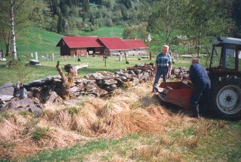 Frå restaureringa av den gamle kyrkjegarden i Bygstad i 1995. Kyrkjegardsmuren vert her retta opp av Mads Laukeland (t.v.) og Gunnar Kleppe. Det sørvestre hjørnet av muren var heilt i ruin då dei byrja. I bakgrunnen ser vi at kyrkjegarden elles er ferdig til innviinga nokre dagar seinare.
