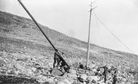 Jakob Kjellevold sitt lag på Aurlandsfjellet. Stolpeskifting i 1936. Arbeidarane er i ferd med å reise ein ny stolpe i steinkaret. Den gamle stolpen er skubba til sides. To mann reiser den nye stolpen og ein mann med spett styrer han på plass i stolpeholet.