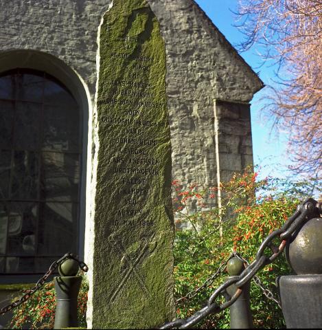 Framsida av minnesteinen med innskrift og våpensymbol. Utananom namna og symbolet krosslagde handvåpen nederst, står det: STENEN * SATTE * VAABENBRØDRE * TIL MINDE * OM (namna) FALDNE * I KAMPEN * VED * ALVØEN * 16. MAI 1808<br />På baksida står: FOR KJÆMPER DER * MED SEIR GIK HJEM * BLEV STENEN SAT * AF NORSKE MÆND