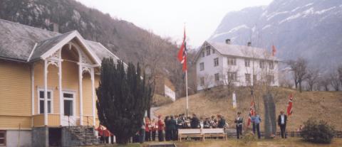 Minnesteinen over dei falne frå Vadheim stod i mange år i hagen ved den gamle dokterbustaden. Her er det samling ved steinen 1. mai 1998 eller 1999. Me ser æresvakt med flagg, musikkorps og blomster ved steinen.